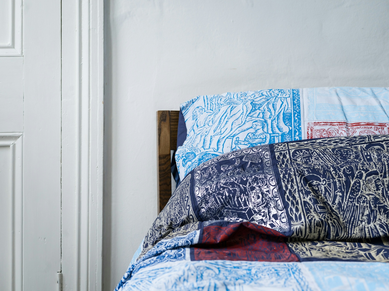Photograph of a bedroom showing the left hand side of the headboard of a double bed, made up with a duvet cover and pillowcases which are made of a patchwork of blue, red, silver and gold screen-printed designs. To the left of the bed is part of a door. 