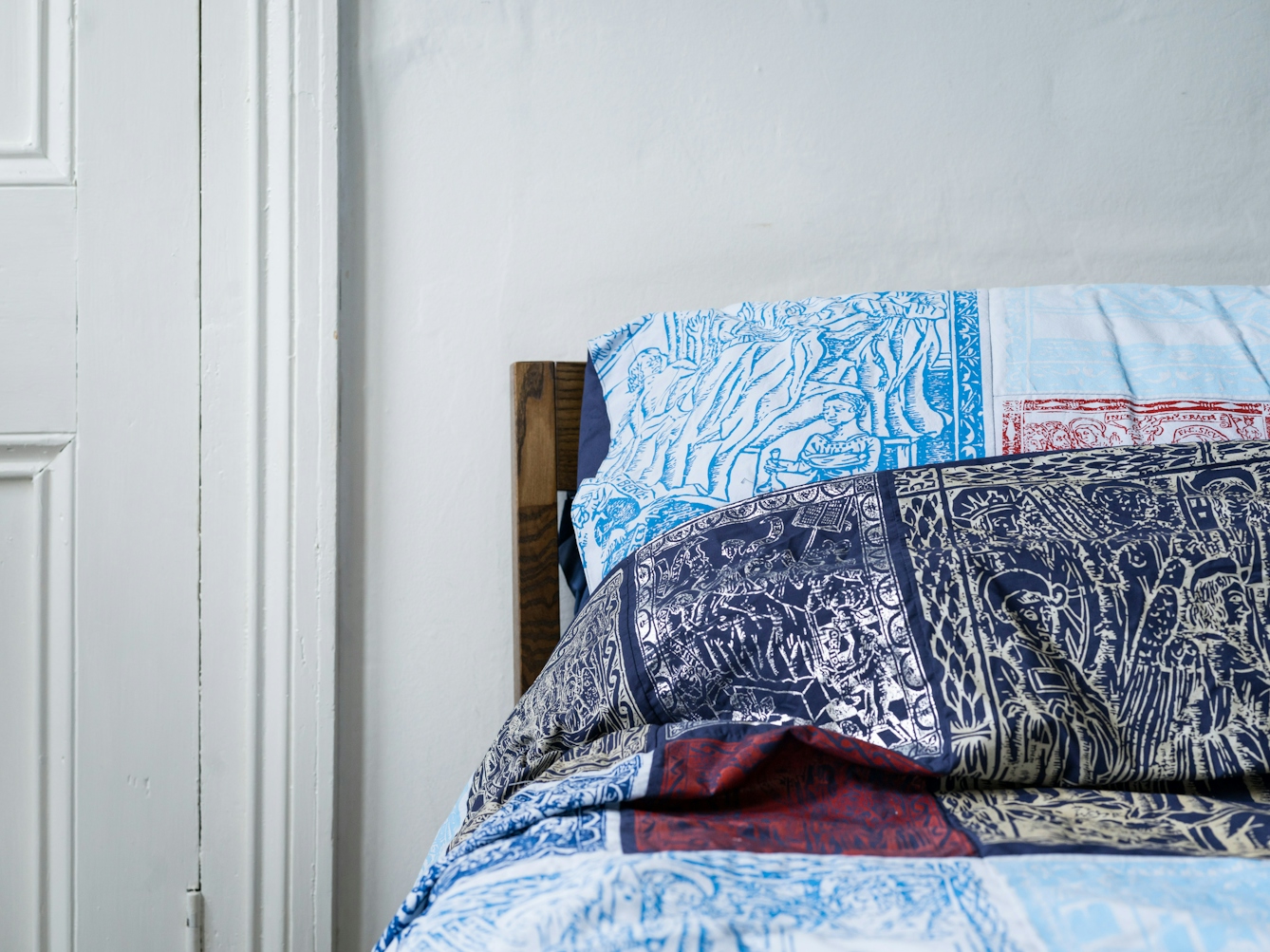 Photograph of a bedroom showing the left hand side of the headboard of a double bed, made up with a duvet cover and pillowcases which are made of a patchwork of blue, red, silver and gold screen-printed designs. To the left of the bed is part of a door.