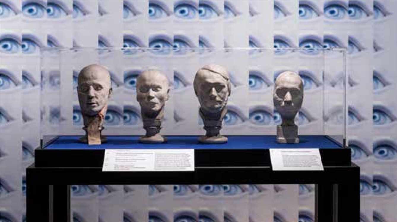 Four whie plaster head busts of men arranged in a row in a glass display cabinet.