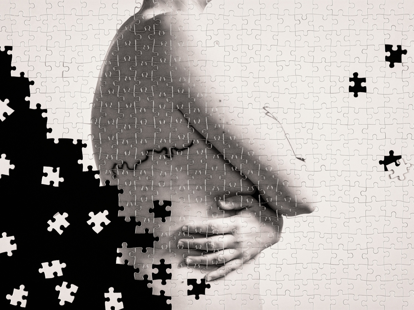 Photograph of a part made jigsaw puzzle containing hundreds of pieces. The jigsaw has been photographed against a black background, with many of the unused pieces scattered around in the black space. The image on the jigsaw is a warmed toned, monotone photograph of a nude woman against a plain light background. The woman is standing showing her right side from the waist to her neck. Her arms are wrapped across her torso, covering her breasts. On her side and back is a tattoo which traces the course of the River Thames. The missing jigsaw pieces create a black void which encroaches from the left and fragments the image of the woman. There are also 3 jigsaw pieces missing or displaced from the solid puzzle on the right side of the image.