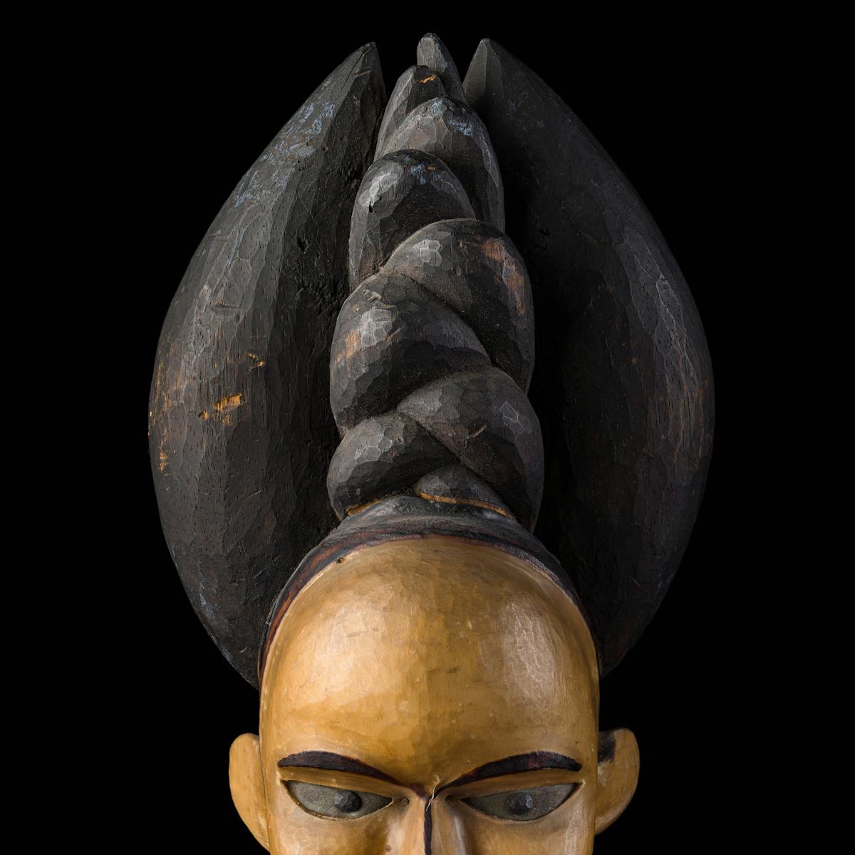 Photograph of the carved wooden head of a woman against a black background. On the woman's head is an elaborate headdress. The head is cropped by the bottom of the frame just below her eyes.