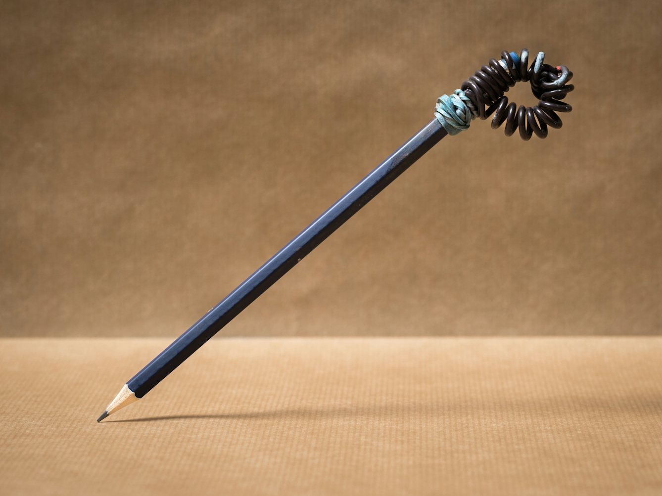 Photograph of a pencil resting at an angle on its sharpened point, the other end suspended in the air. Wrapped around the end of the pencil is a tightly curled wire which makes an open loop. Tye wire is secured to the pencil with a blue elastic band. The background and floor of the image is made with brown parcel paper.