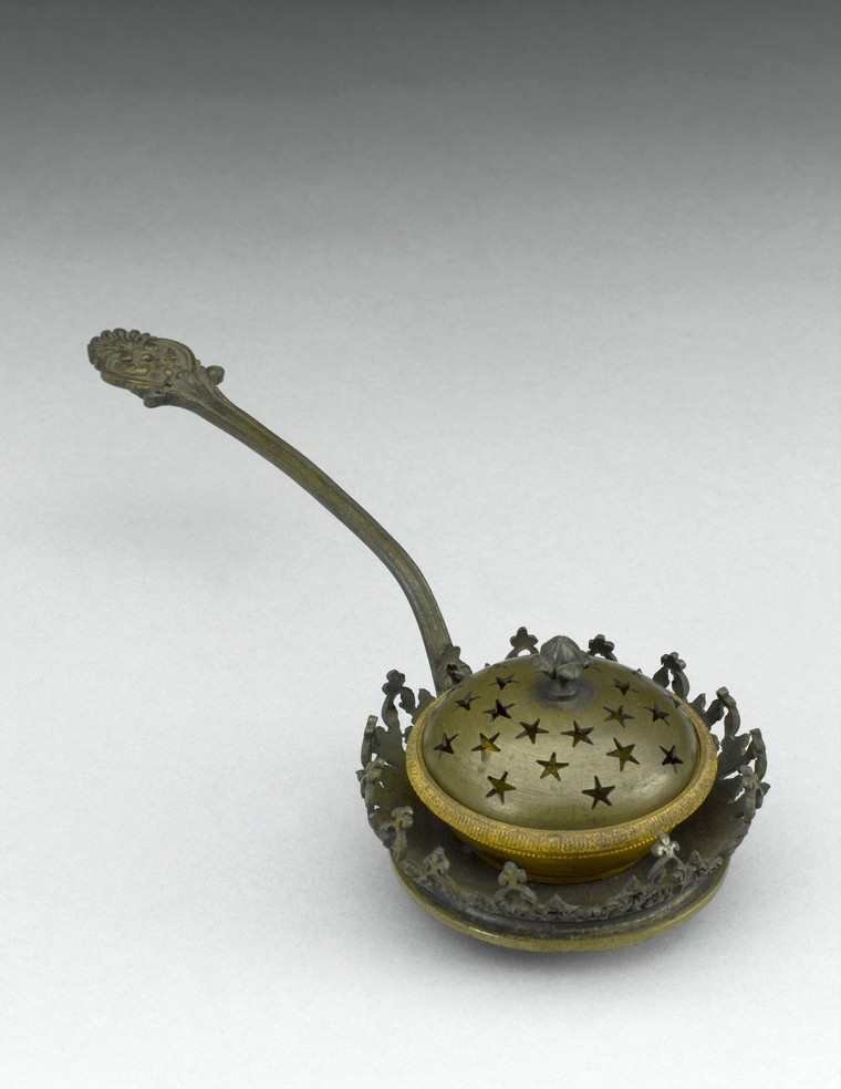 Image of a brass fumigator. Small round container for the incense with star shaped holes, on a short curved handle.