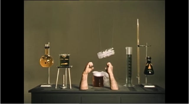 Various scientific flasks and jars are displayed on a flat surface alongside two arms emerging from the table. 