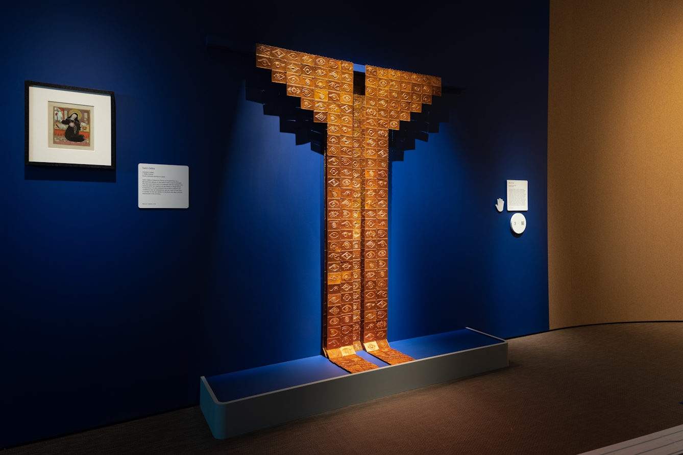 Photograph of an object installation in a gallery setting, spotlit against a dark blue wall. The large object is made out of small copper tiles which have hieroglyphics stamped in relief into their surface. The tiles as a whole create a tall straight form rising up the wall which ends with two wing like shapes either side. Either side of the object are information panels and a small framed print.
