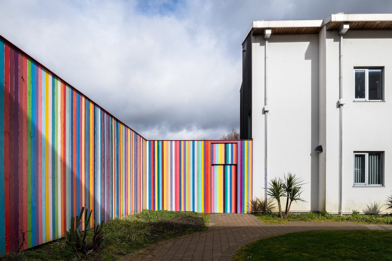 A photograph showing a tall, bright, colourful, striped perimeter fence of which each vertical slat is painted a different colour, consisting mostly of pastel blues, purples, yellows, and pinks. It runs along the left-hand side of the image towards the horizon where it meets a white rectangular looking building with two storeys, and a flat roof. Only one corner of the building is visible with two windows one above the other on separate floors. In the foreground there is short cut green grass, a path leading towards the building. 