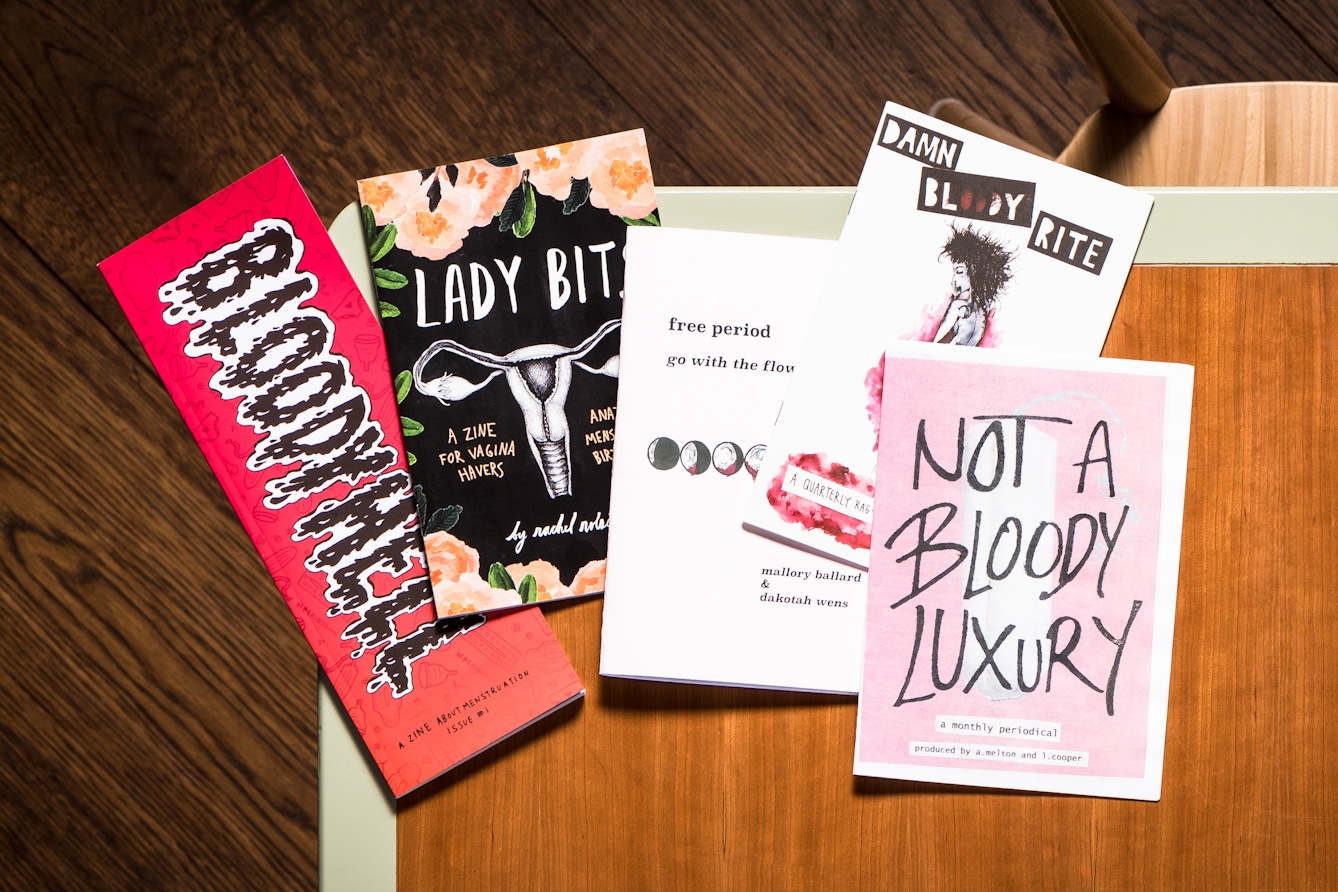 Photograph of a wooden tabletop with a light green edge, from directly above. One the table are 5 zines on the subject of menstruation with titles such as, 'Bloodyhell', 'Lady Bits', 'Damn bloody rite' and 'Not a bloody luxury'.