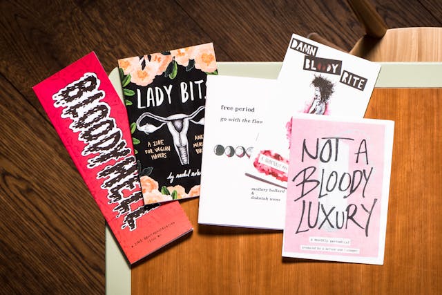 Photograph of a wooden tabletop with a light green edge, from directly above. One the table are 5 zines on the subject of menstruation with titles such as, 