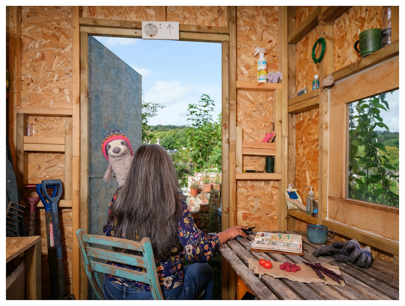 Assisted Self-Portrait of a woman with long hair sat on a wooden chair in an allotment shed with her back to the camera. Peeping over her left shoulder is a grey soft toy sloth, wearing a pink and blue woolly hat, smiling to camera. On the table is an open journal, tomatoes, raspberries and seed pods. In her right hand is the remote trigger that she used to capture the image.