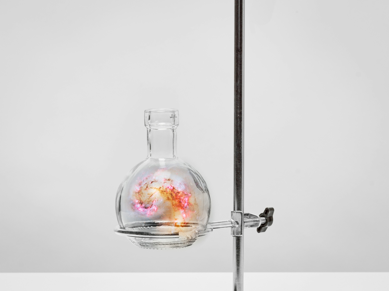 Photograph of a single vertical bar, set against a plain grey background. Clamped to the bar is a horizontal bar and clamp. At the end of the bar is a glass, rounded, flat bottomed flask. Swirling inside the flask is a scene of the galaxy, revealing colourful constellations, swirling stars and gaseous nebula. 