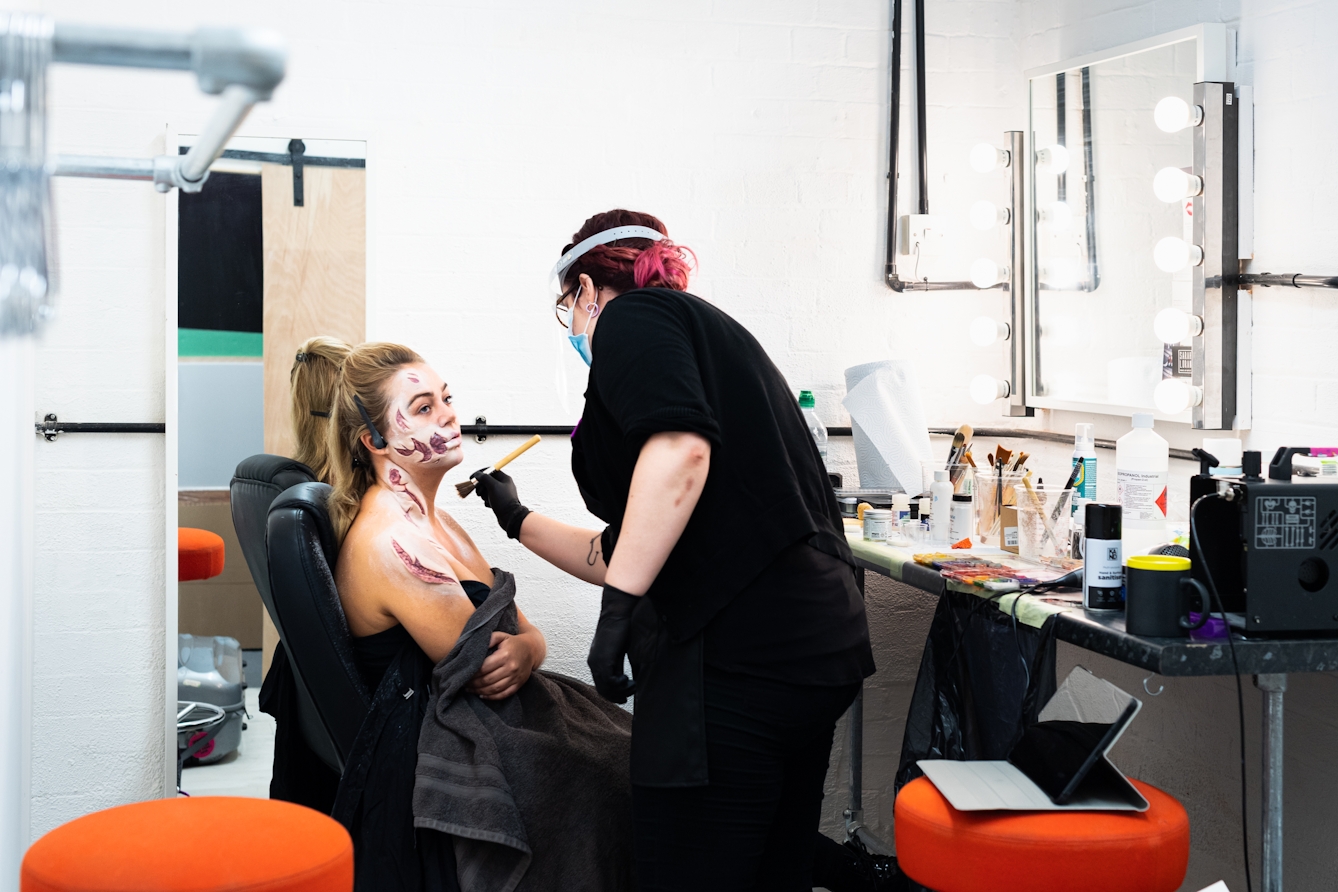 Photograph of a makeup area in a photographic studio with a mirror on the wall surrounded in light bulbs and a high table top beneath. A woman is sitting in a chair wrapped in a black towel with the early stages of prosthetic application on her face and shoulder. A woman in black, wearing a face mask and a visor stands over her with a makeup brush, applying makeup.