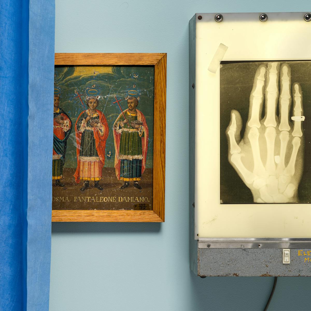 Photograph of an oil painting hung on a light blue wall, depicting 3 saints. The painting is partially hidden behind a blue concertina hospital ward curtain on the left of the frame. To the right is an x-ray lightbox also hung on the wall, with an x-ray of a hand taped to its surface. on the little finger of the hand you can see the outline of a ring. 