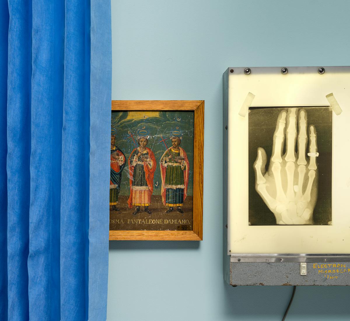 Photograph of an oil painting hung on a light blue wall, depicting 3 saints. The painting is partially hidden behind a blue concertina hospital ward curtain on the left of the frame. To the right is an x-ray lightbox also hung on the wall, with an x-ray of a hand taped to its surface. on the little finger of the hand you can see the outline of a ring. 