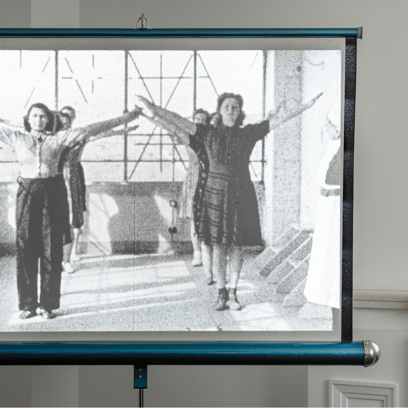 Photograph of a Hunter Starlight projector screen in the corner of room by a window recess. On the screen is an still from a film, showing two lines of expectant mothers with their arms raised as part of an excessive class. To the right of the frame a nurse is just visible.