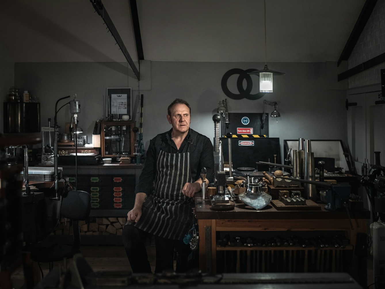 Photograph.  Portrait of a man sitting on a stool in his workshop.  His arm is leaning on a wooden workbench on which there are tools including a row of hammers and various other metal working tools. 