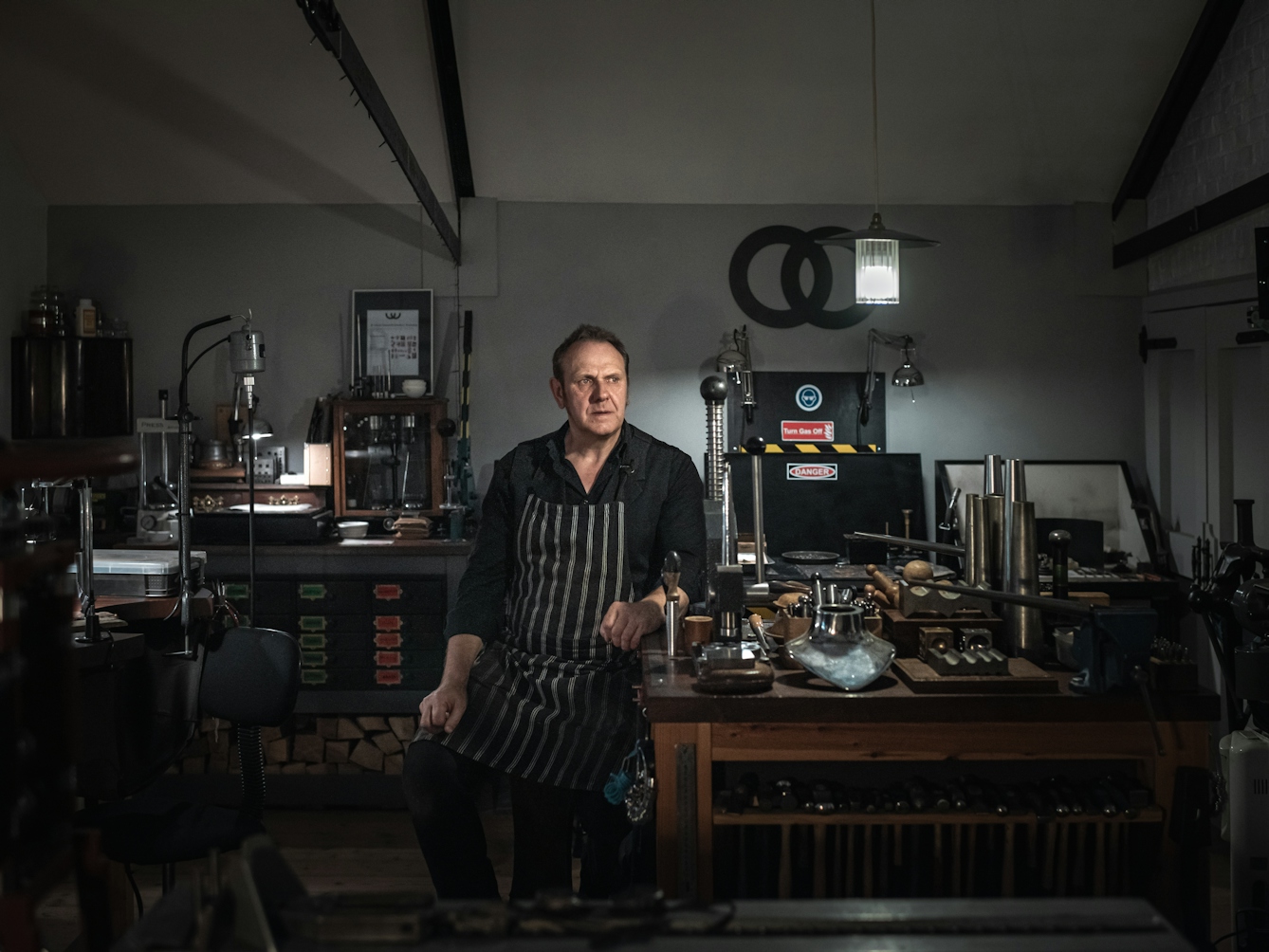 Photograph. Portrait of a man sitting on a stool in his workshop. His arm is leaning on a wooden workbench on which there are tools including a row of hammers and various other metal working tools.