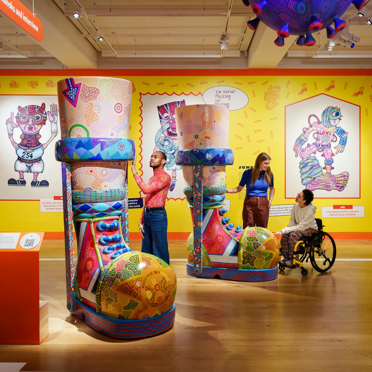 Photograph of a colourful exhibition space with three individuals exploring a large sculpture of a pair of boots with callipers. The sculpture is covered in colourful patterns and towers above their heads. On the wall in the background is a bright yellow mural containing drawings and text. 