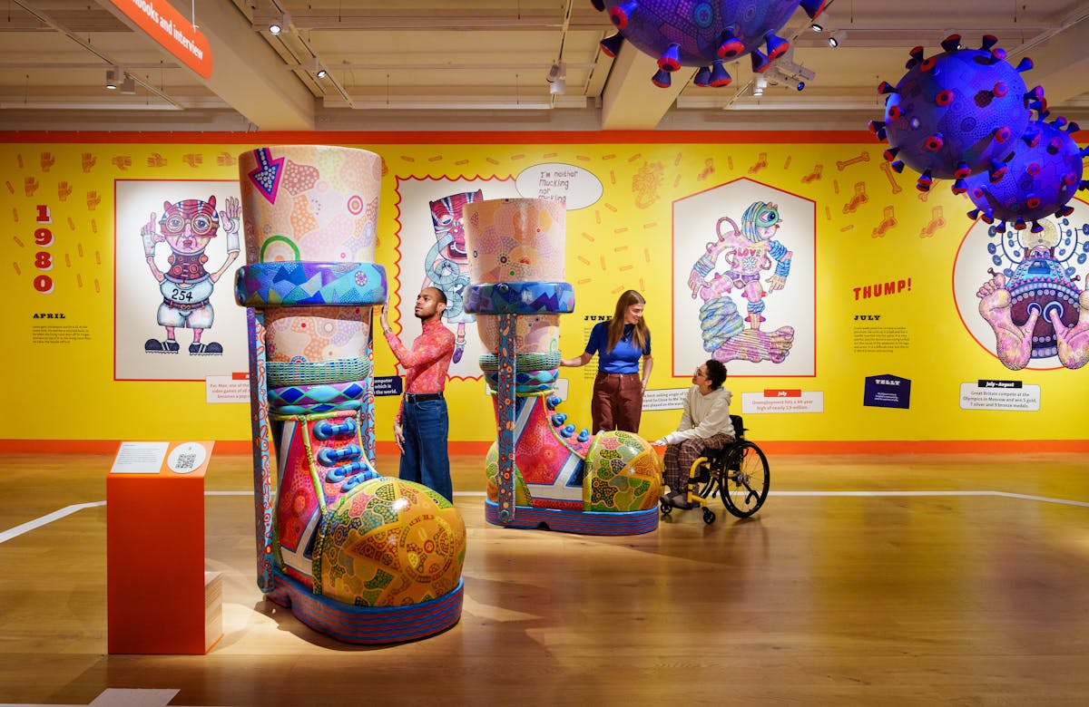 Photograph of a colourful exhibition space with three individuals exploring a large sculpture of a pair of boots with callipers. The sculpture is covered in colourful patterns and towers above their heads. On the wall in the background is a bright yellow mural containing drawings and text. 