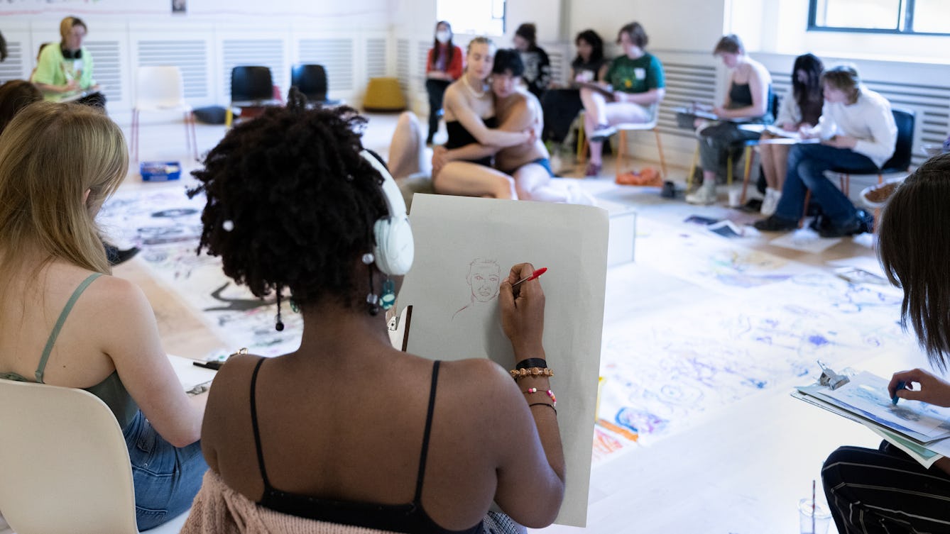 A life drawing workshop showing a young person sketching a pen drawing of figures who can be seen posing in the background.