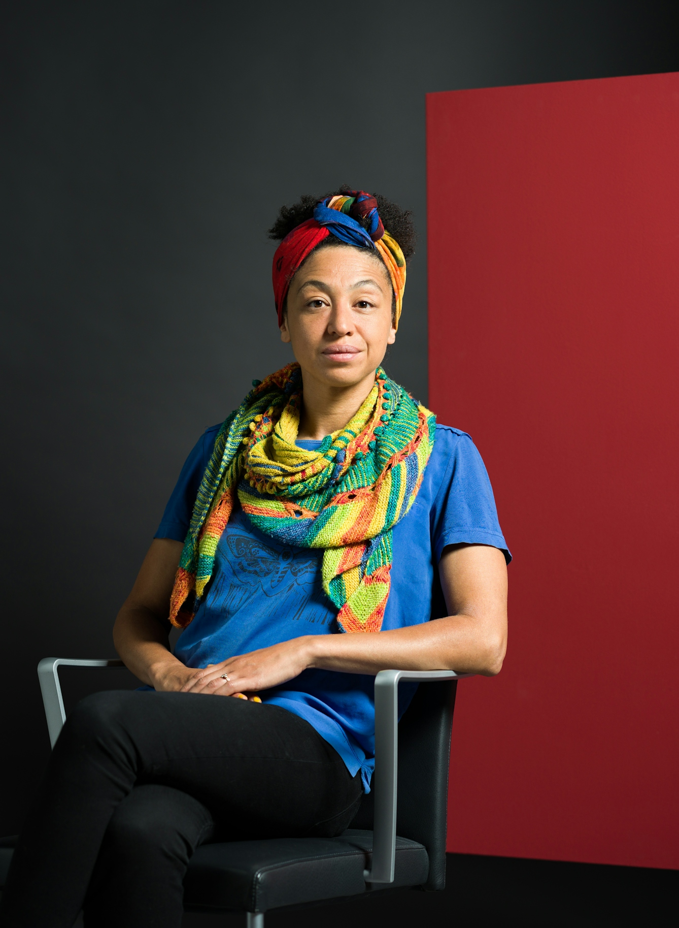 Colour photograph three-quarter length portrait of Charmaine Wombwell, who is seated in a chair. Charmaine is has the beginnings of a smile around her eyes and the corners of her mouth. She is black and has curly hair swept up behind a colourful wrap, knotted neatly at the top of her head. She wears a blue t-shirt and dark trousers, and a scarf patterned with lime green, forest green and orange. 