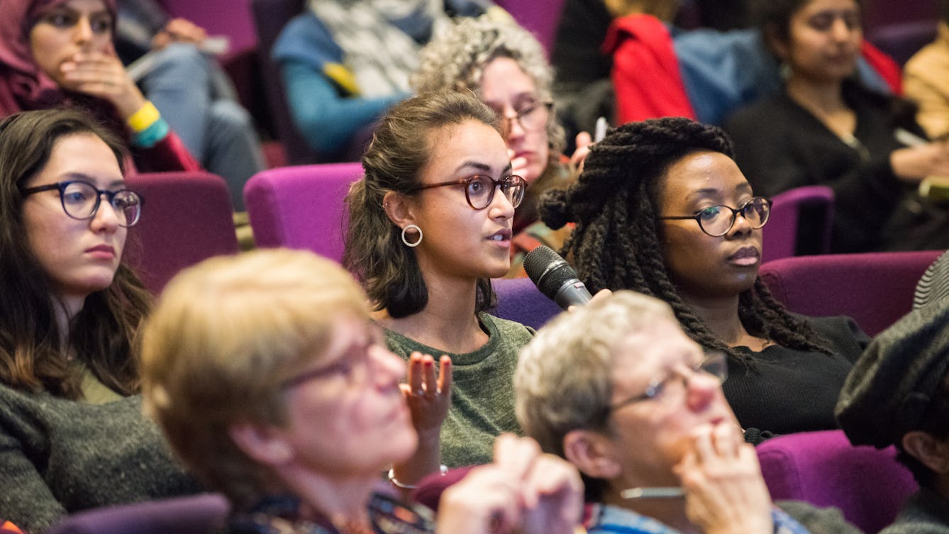 People sitting in rows of purple seating in the auditorium at Wellcome Collection, a member of the audience is speaking into a handheld microphone and gesturing with their other hand.