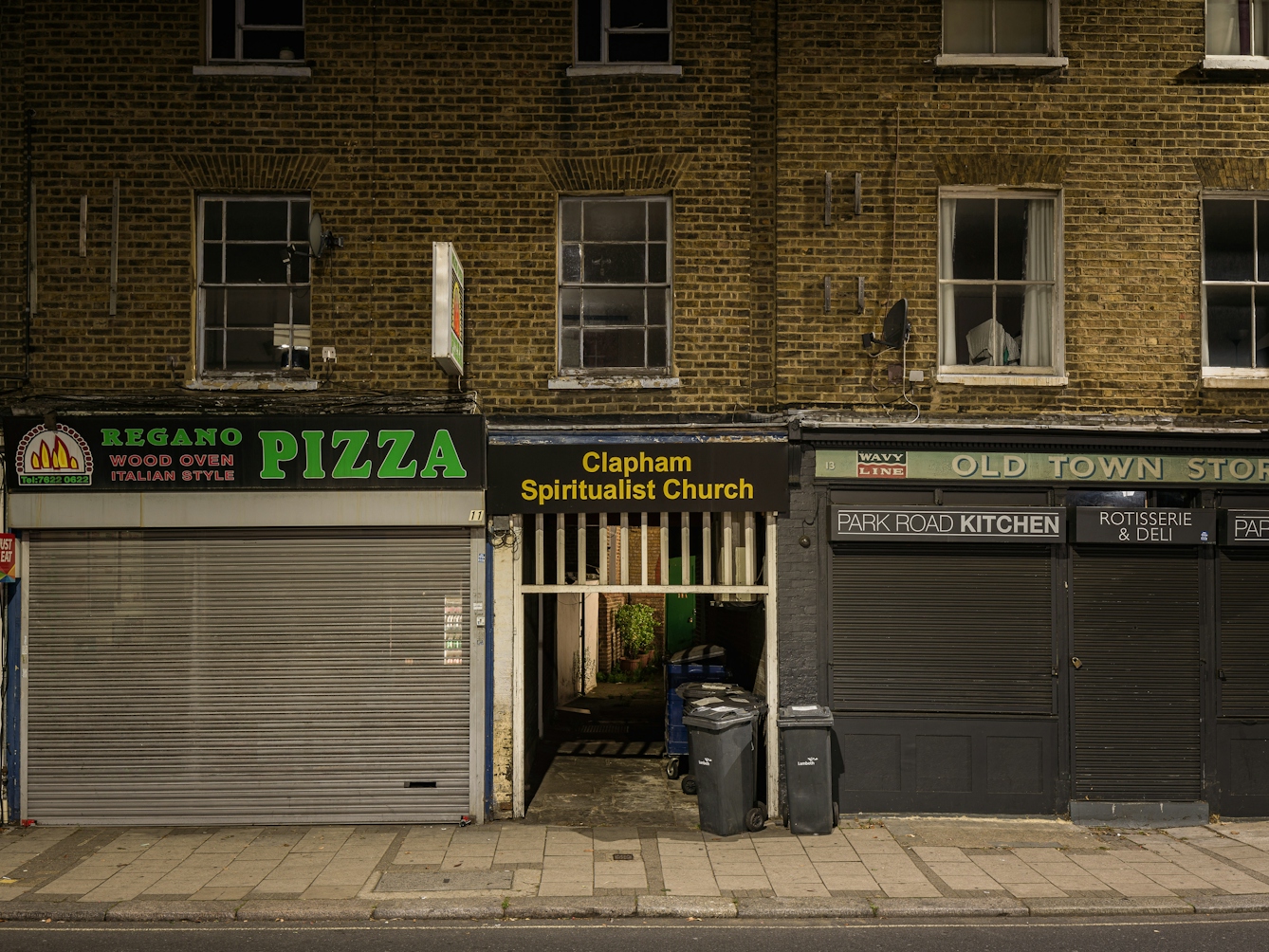 Photograph of the tunnelled entrance to Clapham Spiritualist Church at night.  The church is concealed behind a terraced building, the ground floor of which are commercial properties.  The tunnel has a sign above it that reads 'Clapham Spiritualist Church' in yellow lettering on a black background.  To the left of the entrance is a Pizza takeaway shop, and to the right is the 'Old Town Store' Rotisserie & Deli Shop.
