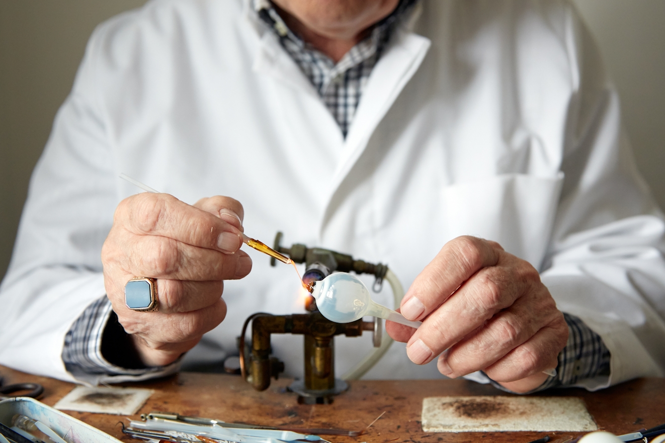 Photograph of the hands of a man in a white lab coat, holding a tube of white glass with a sphere at one end, to the flame of a gas torch. In one hand is a length of brown coloured glass tube which is being used to create a coloured disk onto the surface of the sphere. Next to him on the workbench are thin tools and more lengths of glass tubing.