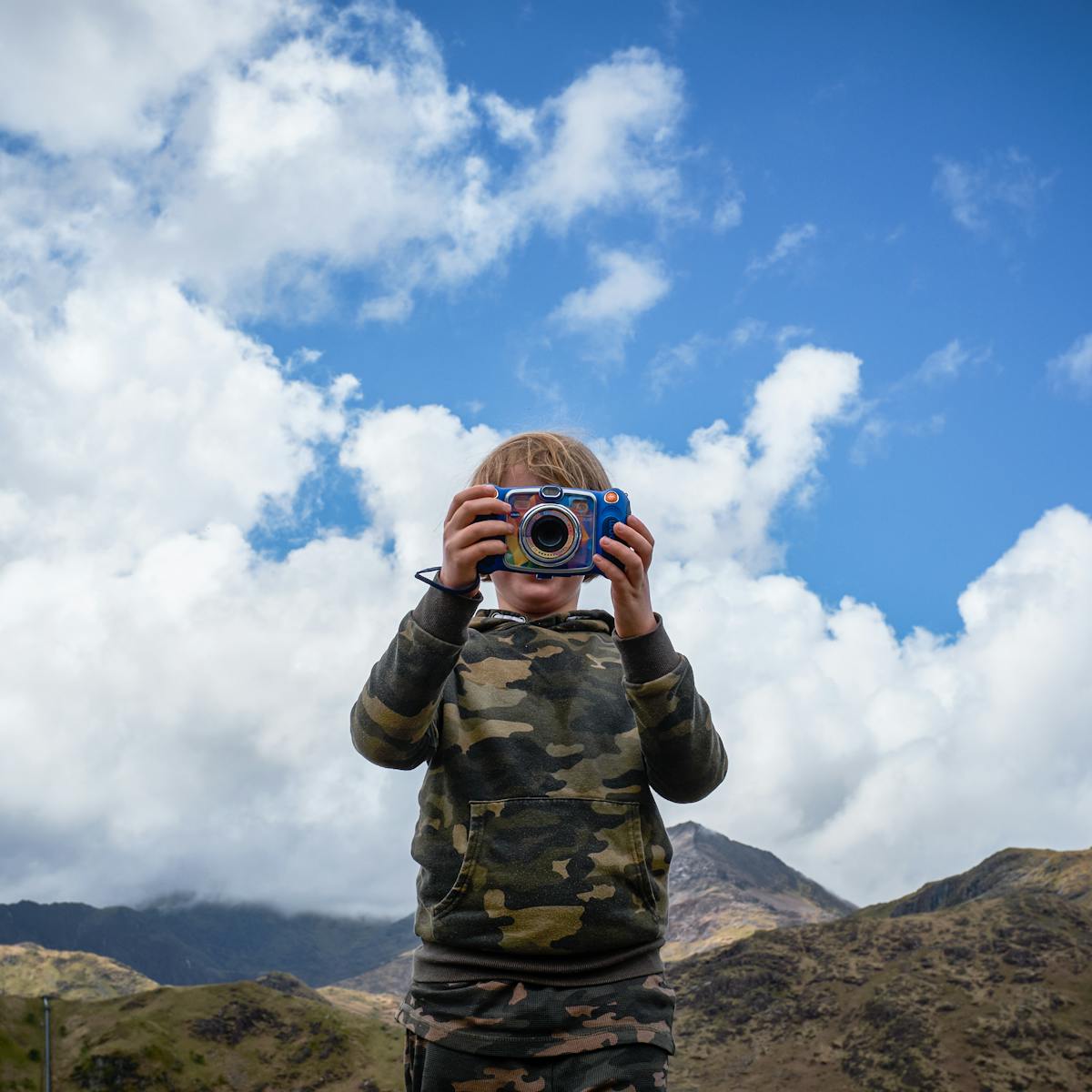 Photograph of a young child with blonde hair holding up a colourful camera. The camera obscures their face and they are wearing a camouflage jumper and tracksuit bottoms. There are some mountains behind them and the sky is blue and cloudy. 