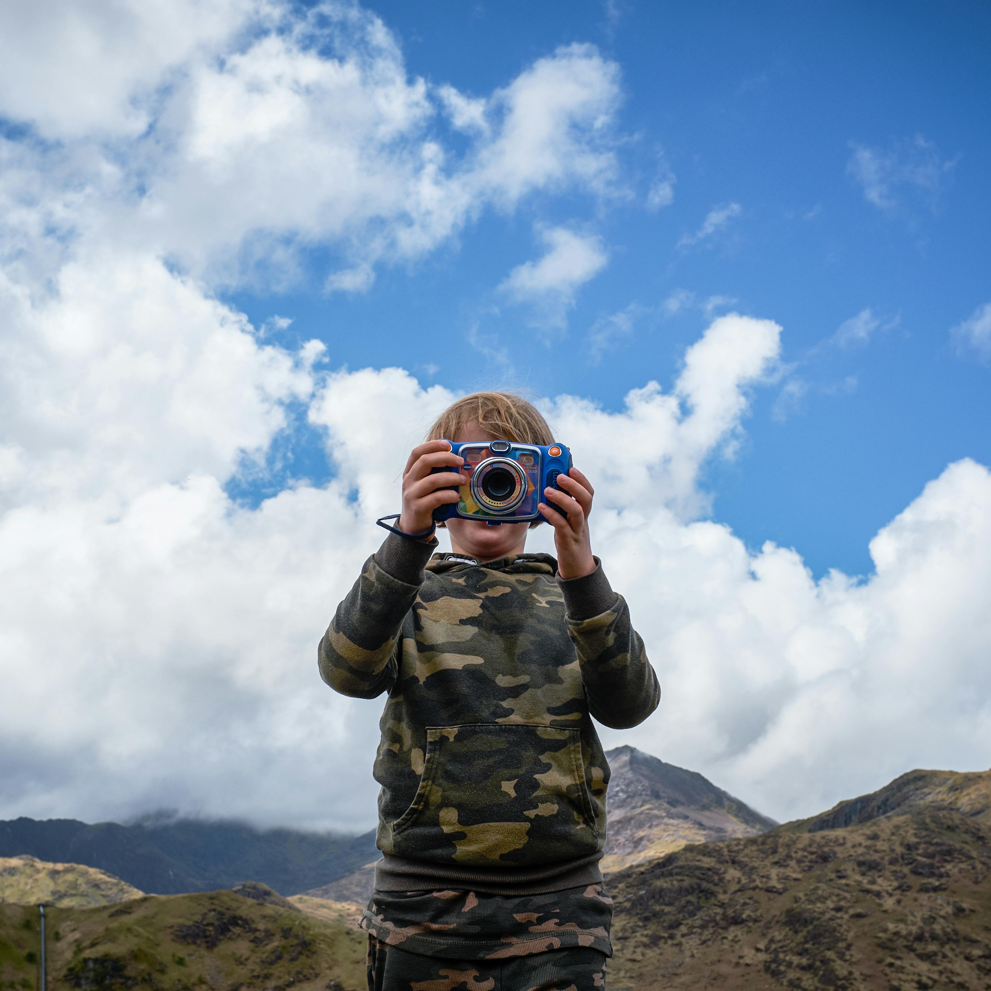 Photograph of a young child with blonde hair holding up a colourful camera. The camera obscures their face and they are wearing a camouflage jumper and tracksuit bottoms. There are some mountains behind them and the sky is blue and cloudy.