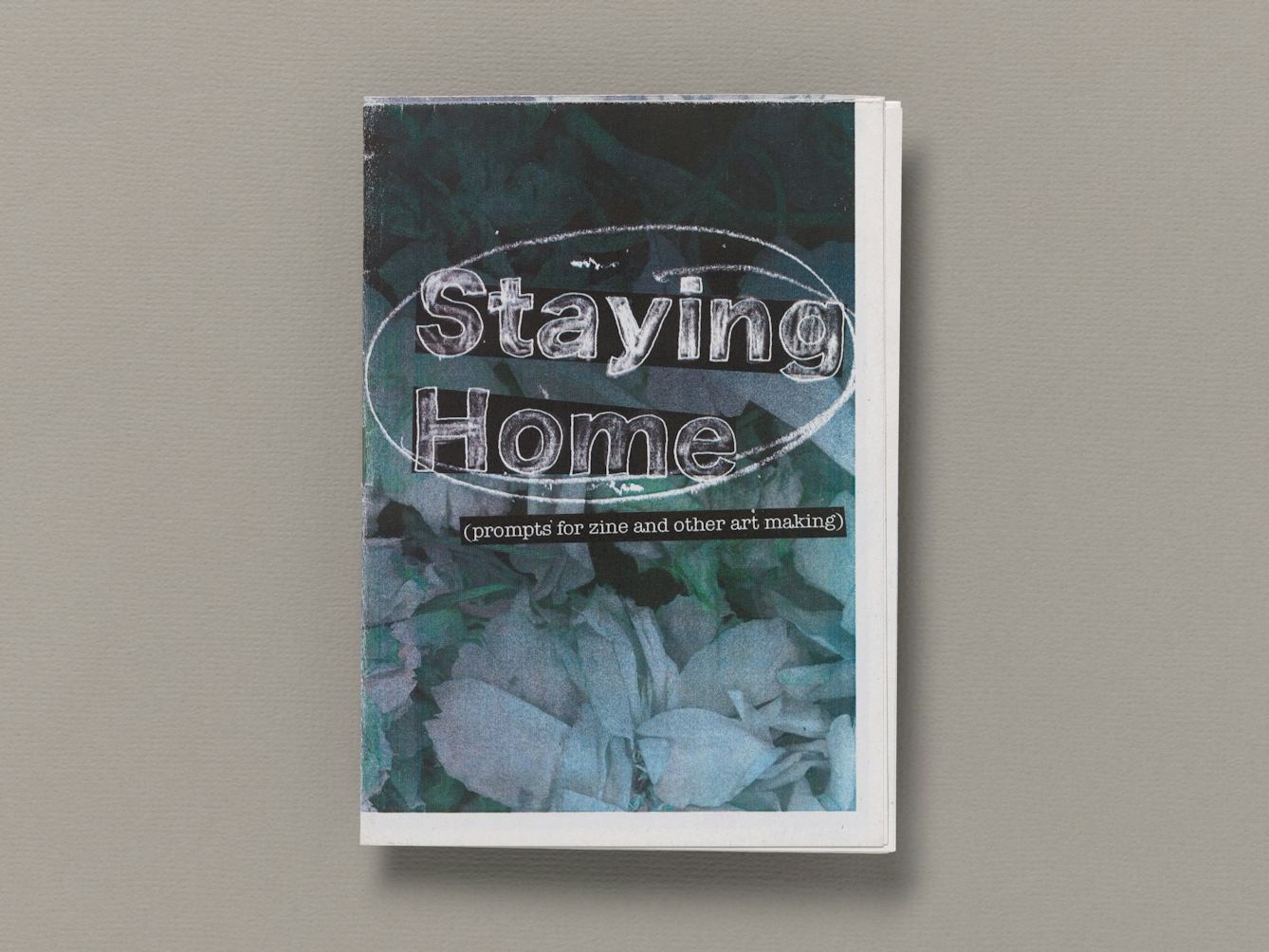 The front cover of the zine "Staying home (prompts for zine and other art making). by Vicky Stevenson. The words "Saying home are written in large outline  in the middle third of the cover and roughly filled in with a white chalky efet against a black background. The words are roughly circled in the same chalk like marker. Imediately below this is the subtitle in brackets in white typeset in a narrow black strip. The background of the whole page is an abstract pattern of pale white shapes tinged with bluish green against a black background.