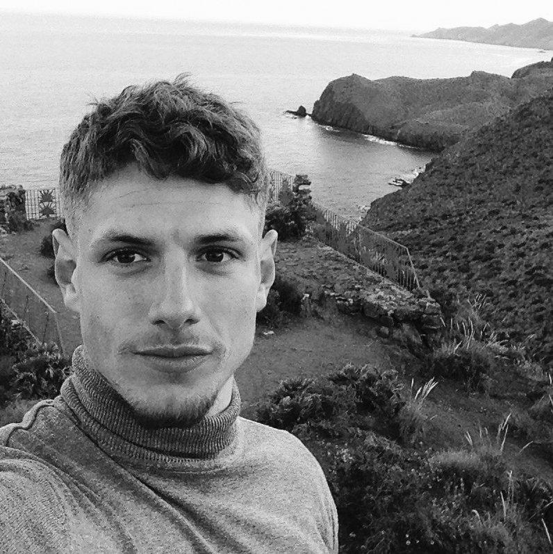 Head and shoulders photograph of a man with shortish hair and a tidy beard and moustache. In the background, green hills fall to the sea along a coastline. 