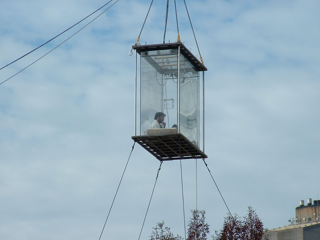Colour photograph showing a man sitting inside a clear box suspended in the air.
