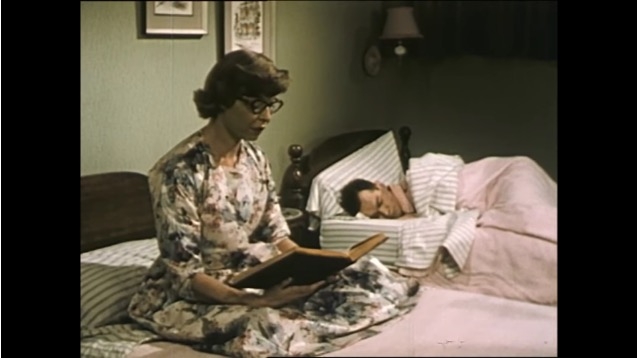 Still image from film of a man asleep in bed while a woman reads from a book