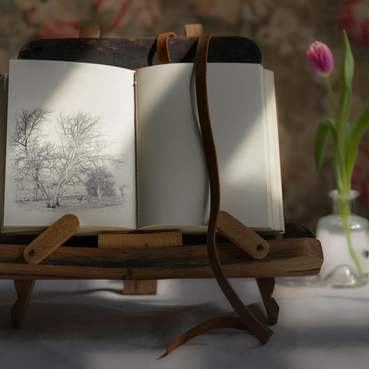 Photograph of a notebook on a wooden book stand, lit by a window from the right, casting a shadow on half of the page. On the left page there is a sketch of a landscape with trees and a bench. To the right of the book a single tulip in a small glass vase.