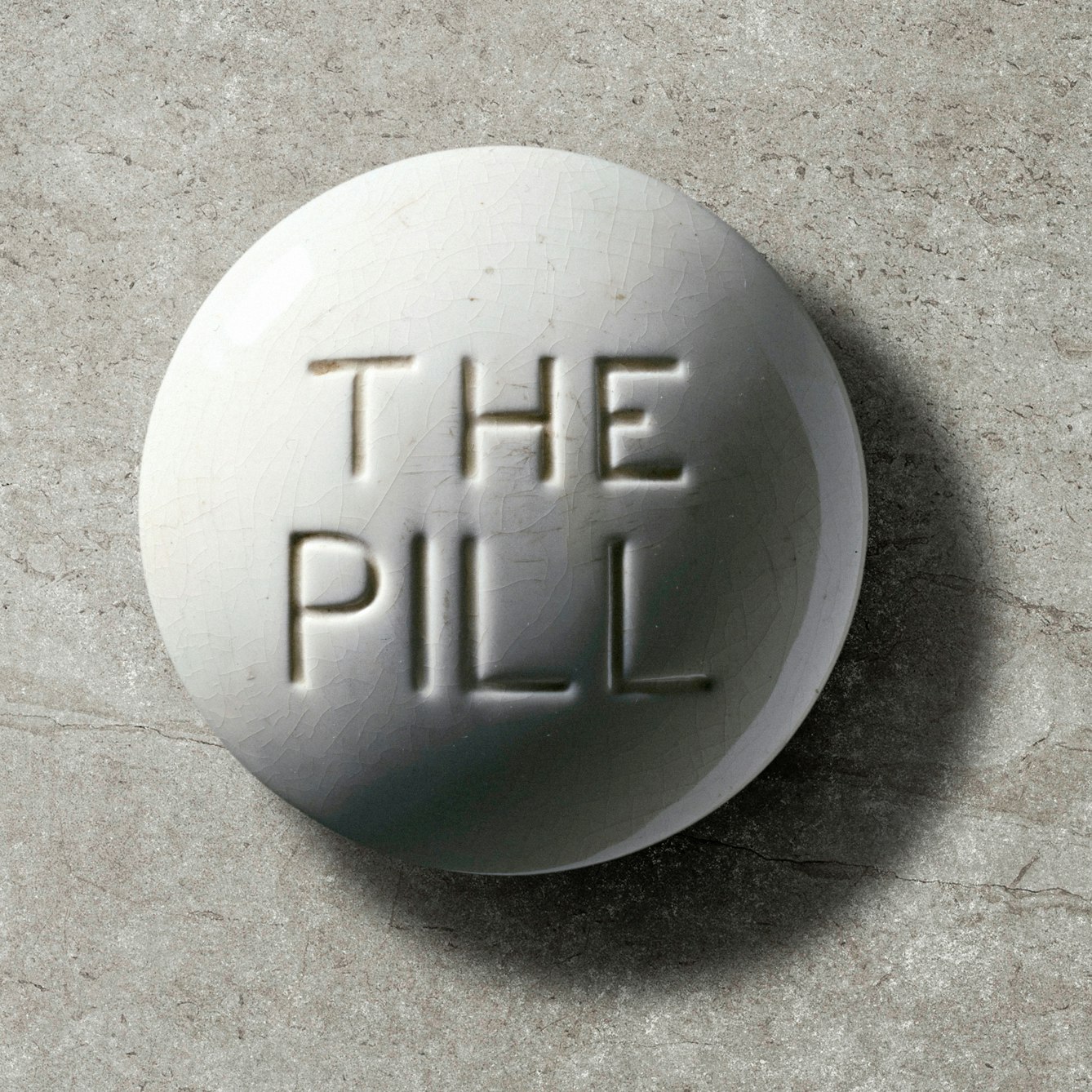 Photograph of a contraceptive pill model, Europe, c. 1970. White crackled glaze, with debossed "THE PILL" text in centre, on a grey concrete textured background.