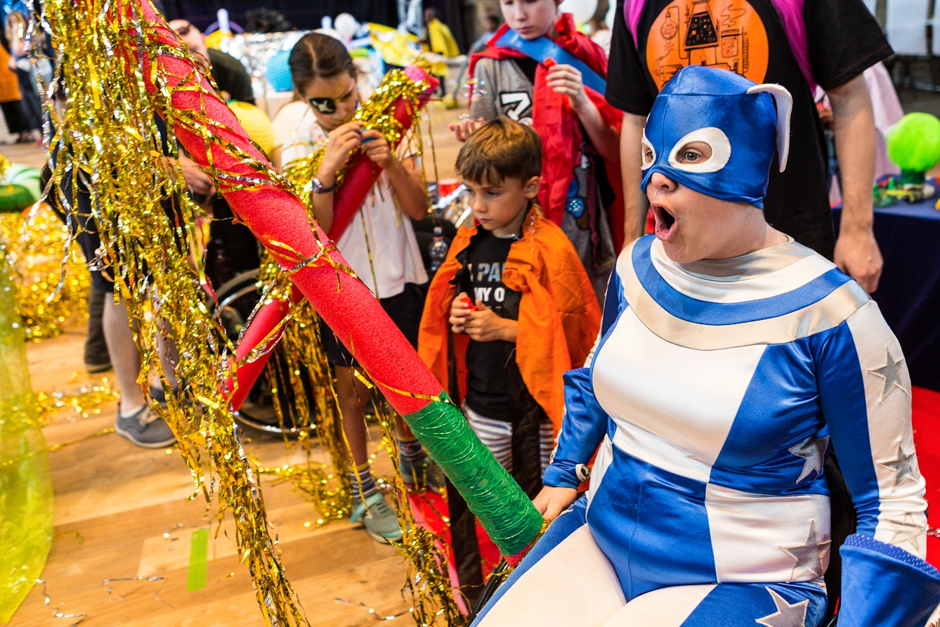 Photograph at an event showing a woman wearing a blue and white superhero style outfit, with her mouth wide open. She is accompanied by children involved in the activity.