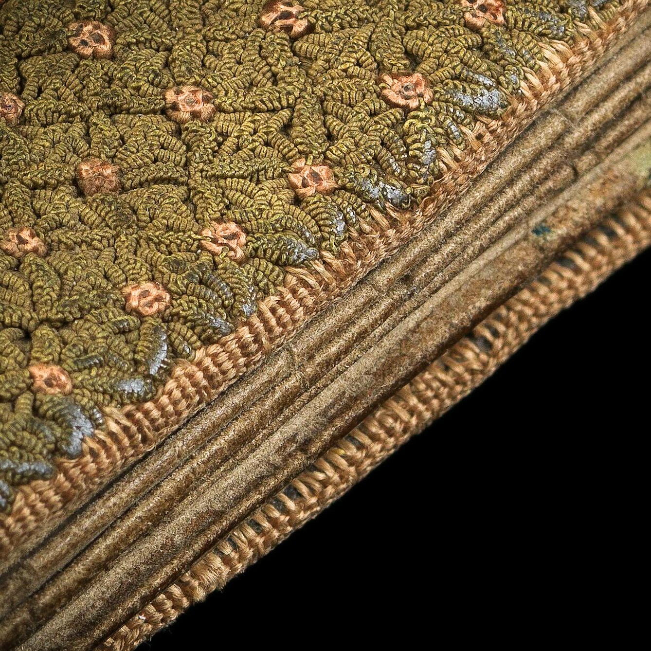 Photographic detail of a fragile folding almanac from the 15th century. The almanac is spotlit on a black background. It is photographed extremely close up so that the threads which make up its intricate woven green and pink fabric cover can be clearly seen.