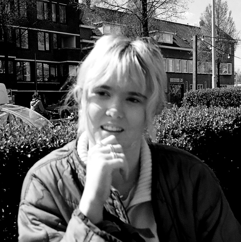 Black and white photograph of a young blonde woman standing in front of a hedge with an apartment building beyond. She looks like she is in conversation and has her right hand raised to her chin