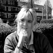 Black and white photograph of a young blonde woman standing in front of a hedge with an apartment building beyond. She looks like she is in conversation and has her right hand raised to her chin