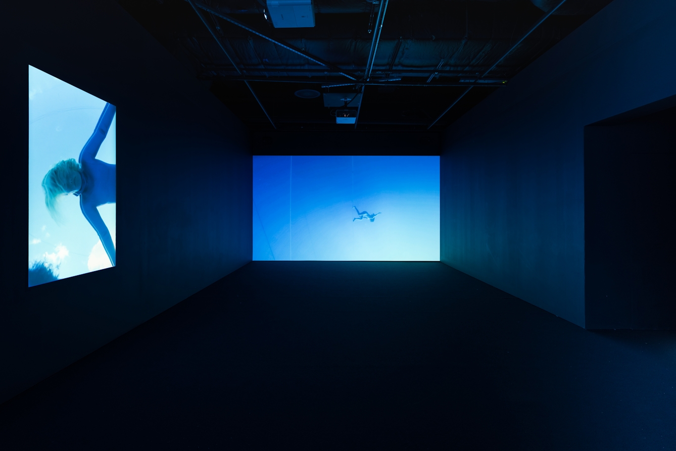 Photograph of a dark gallery with a large projection showing figures floating in water against one wall, which casts blue light into the room.