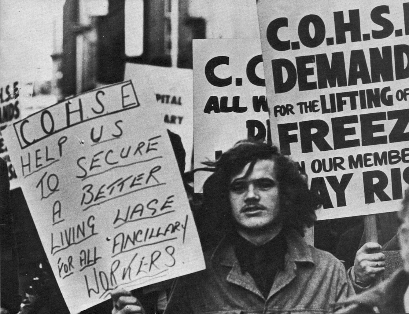 Black and white photograph of a protest with banners reading "COHSE help us to secure a better living wage for all ancillary workers" and "COHSE demand for the lifting of freeze on our members' pay rise"
