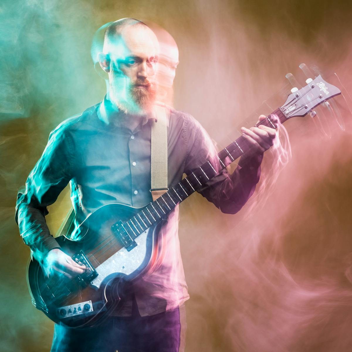 Photograph of a young man playing a bass guitar against a dark yellow background. The slow exposure of the camera causes his movements to be traced across the image as a blur. He is surrounded by wisps of smoke which are coloured cyan on the left of the frame and a pink on the right.