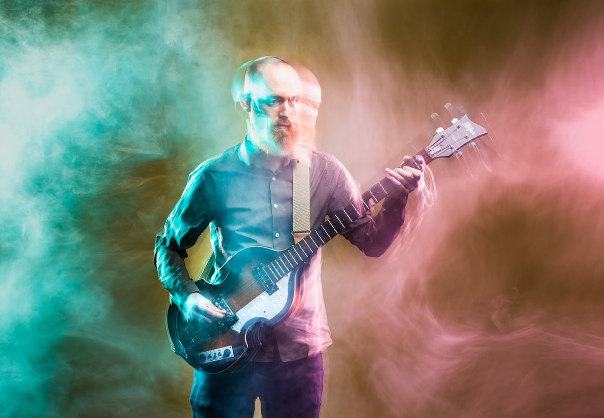 Photograph of a young man playing a bass guitar against a dark yellow background. The slow exposure of the camera causes his movements to be traced across the image as a blur. He is surrounded by wisps of smoke which are coloured cyan on the left of the frame and a pink on the right.