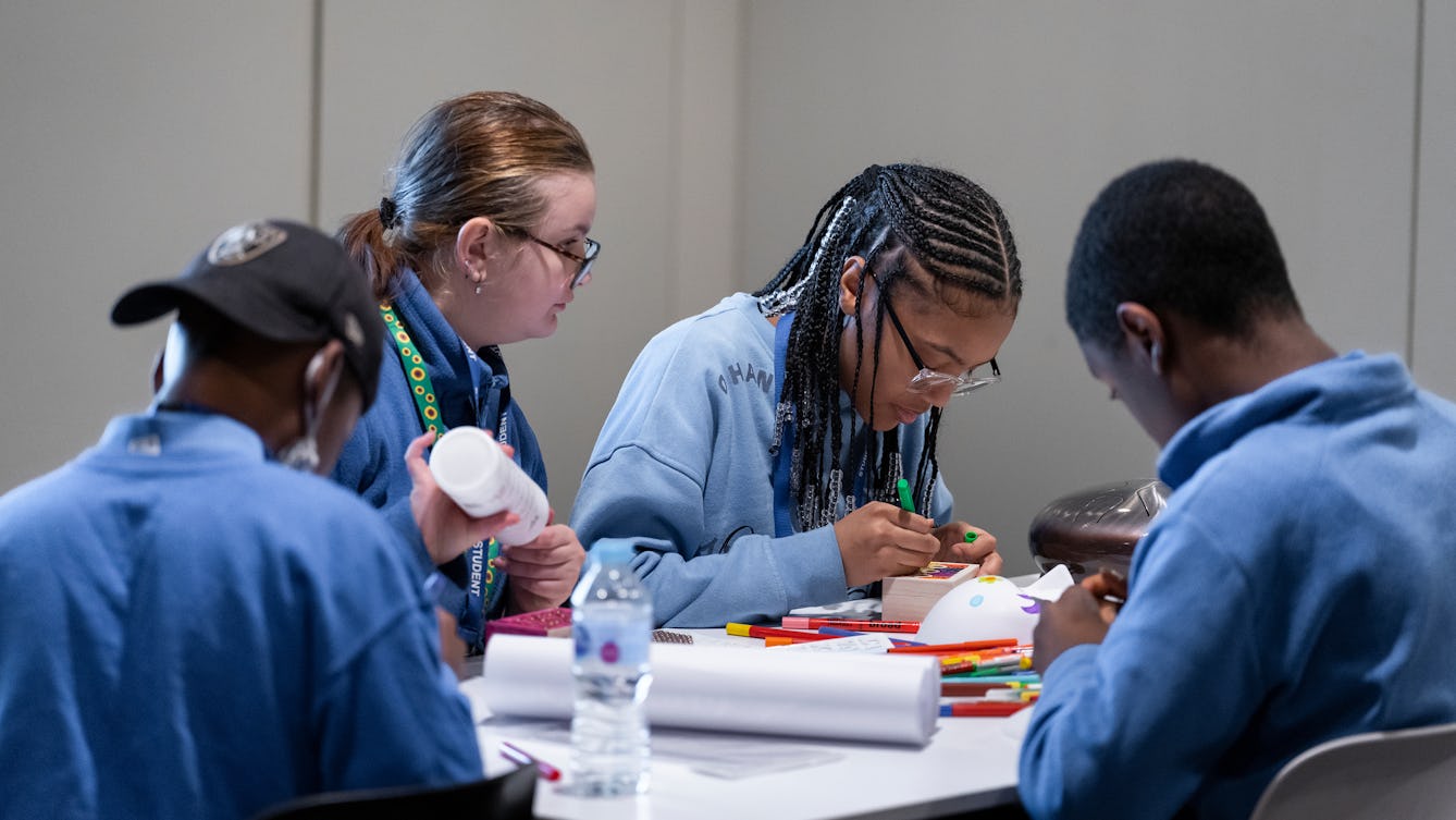 Four young people in a blue school uniform sitting around a table with pens and paper engaged in a creative workshop activity. 
