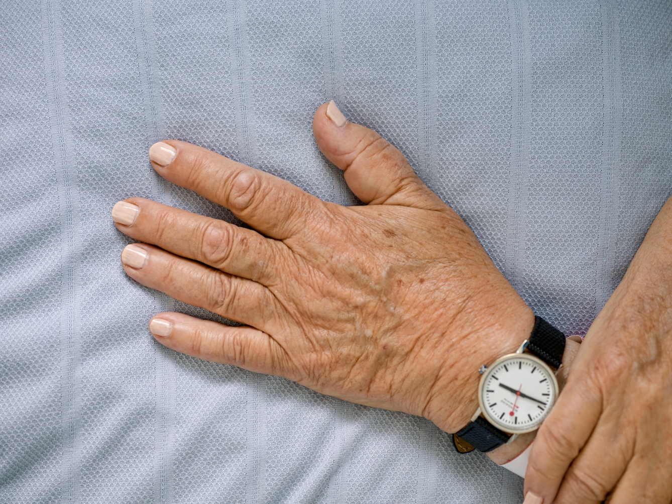 Photograph of the hands of a patient with light pink painted fingernails, resting on a light blue hospital bedsheet.