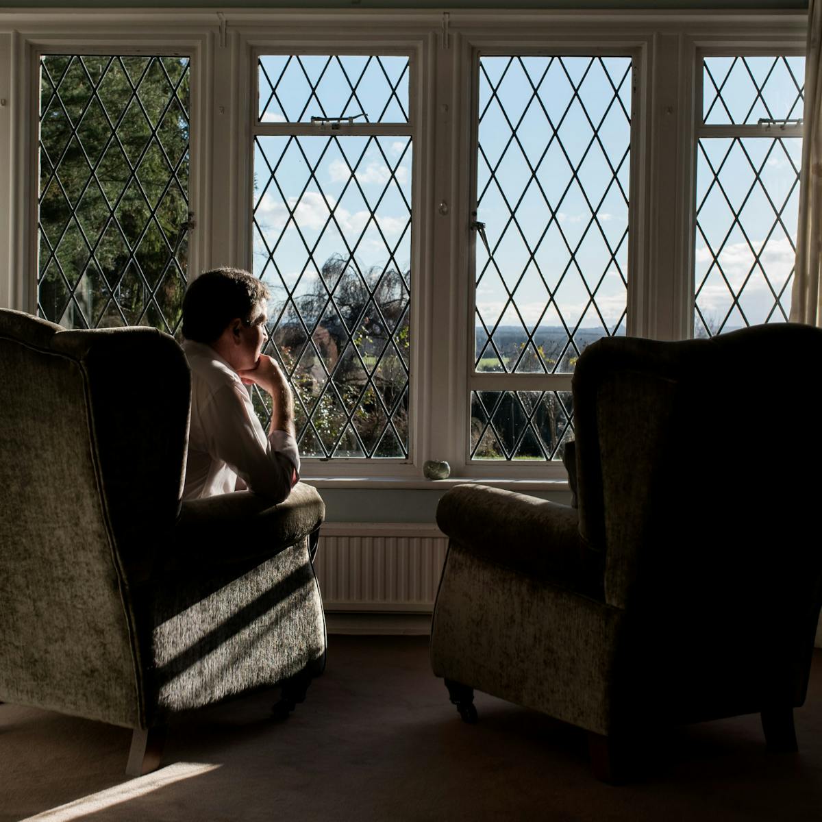 Photograph of a man sat in a living room armchair looking out of a window, his chin resting on his hand. To his right is an identical armchair which is empty,