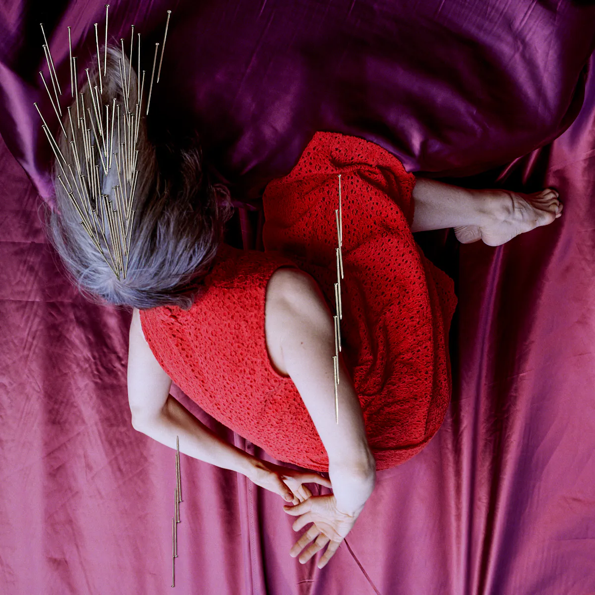 Artwork created with a colour photographic print of a female figure in a bright red dress, set against a purple and blue draped silk background. She is curled up in the frame with her hands loosely clasped behind her back, caught as if in mid fall, hair blown upwards. Her face is obscured by her hair. Her body is targeted by groups of dress pins, laid on top of the photographic print. The pins are arranged as if they are a flight of arrows directed at her body. Two groups attack her arms from underneath and above. Another final group descend from above towards the crown of her head.