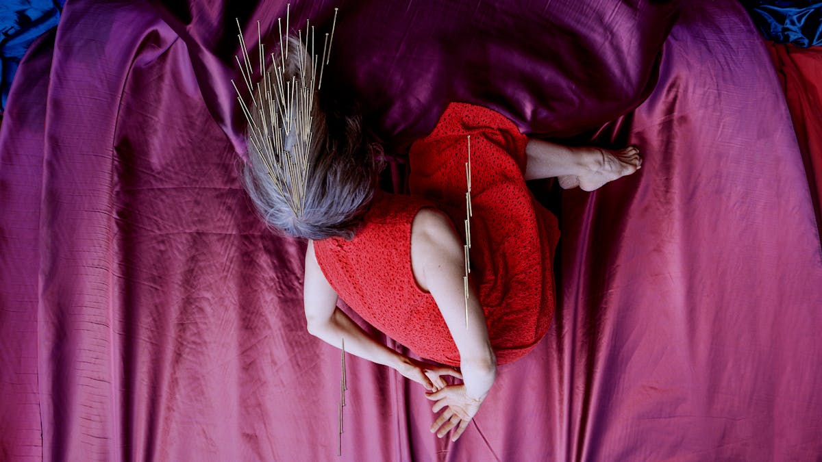 Artwork created with a colour photographic print of a female figure in a bright red dress, set against a purple and blue draped silk background. She is curled up in the frame with her hands loosely clasped behind her back, caught as if in mid fall, hair blown upwards. Her face is obscured by her hair. Her body is targeted by groups of dress pins, laid on top of the photographic print. The pins are arranged as if they are a flight of arrows directed at her body. Two groups attack her arms from underneath and above. Another final group descend from above towards the crown of her head.