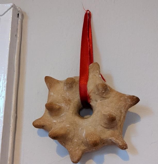 Photograph of a witch cake in a star shape hanging on a red ribbon. The ribbon is attached to a nail on a white wall. 