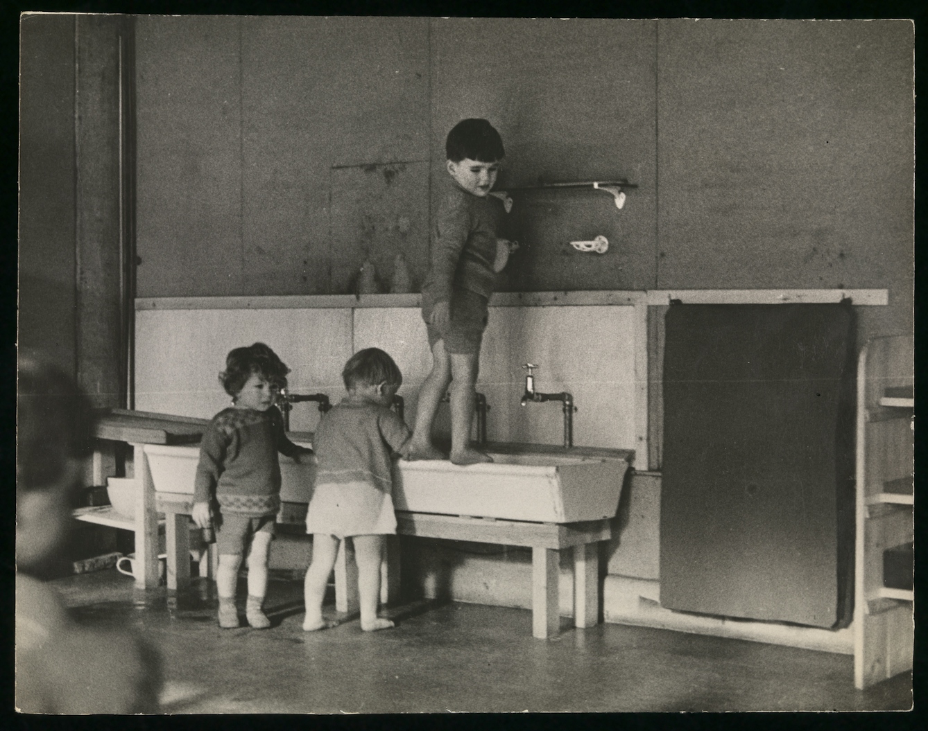 Black and white photograph showing three toddlers at a long, low wash basin.
